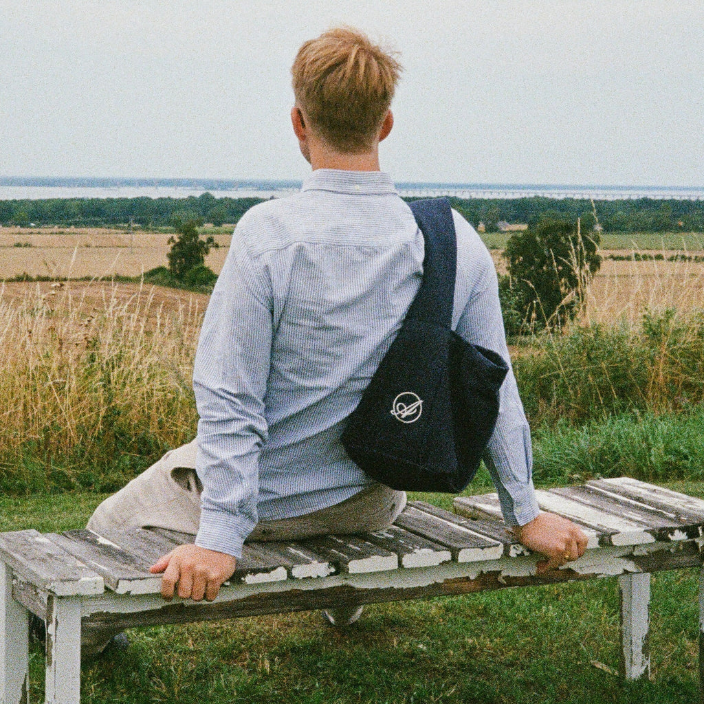 A man sitting on a bench looking over fields, with the navy Shadow Extra Bag across his back
