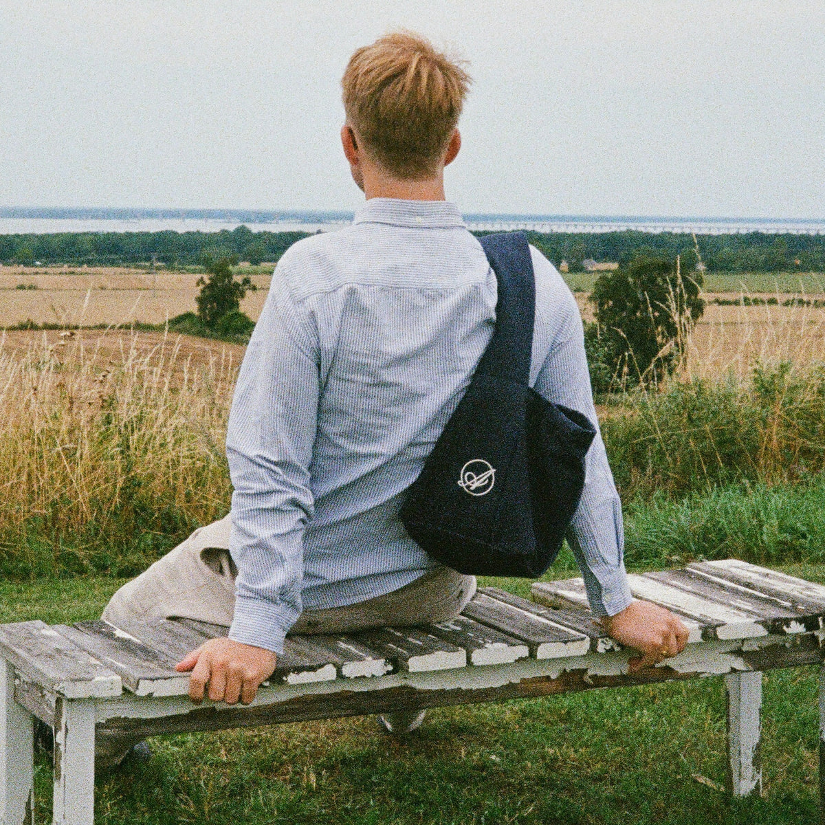 A man sitting on a bench looking over fields, with the navy Shadow Extra Bag across his back