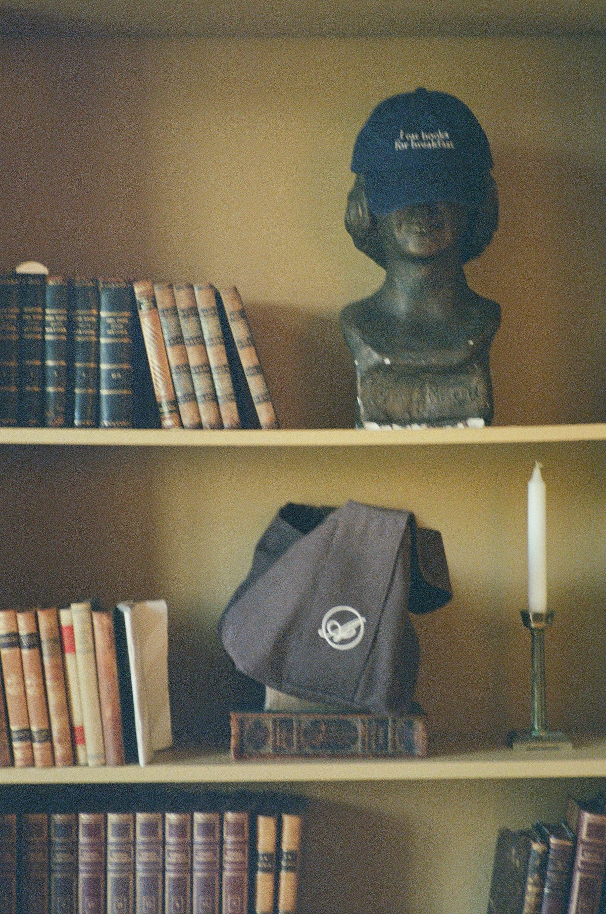 A bookshelf with old leather-bound books, a bust wearing a navy Abelo cap and a dark brown Abelo bag resting on a book