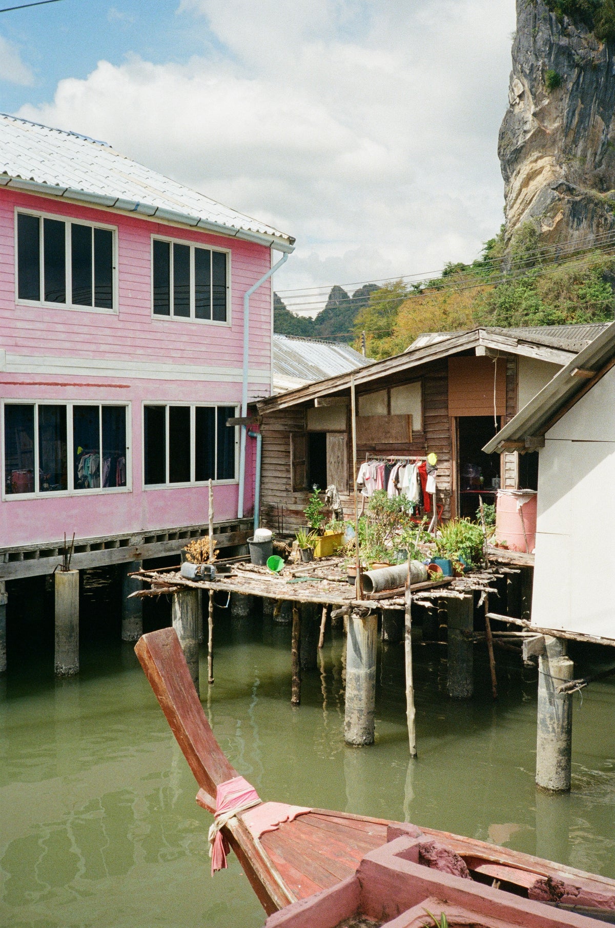 Pink and white stilt houses above green water, with a longtail boat moored below, in Phang Nga Bay