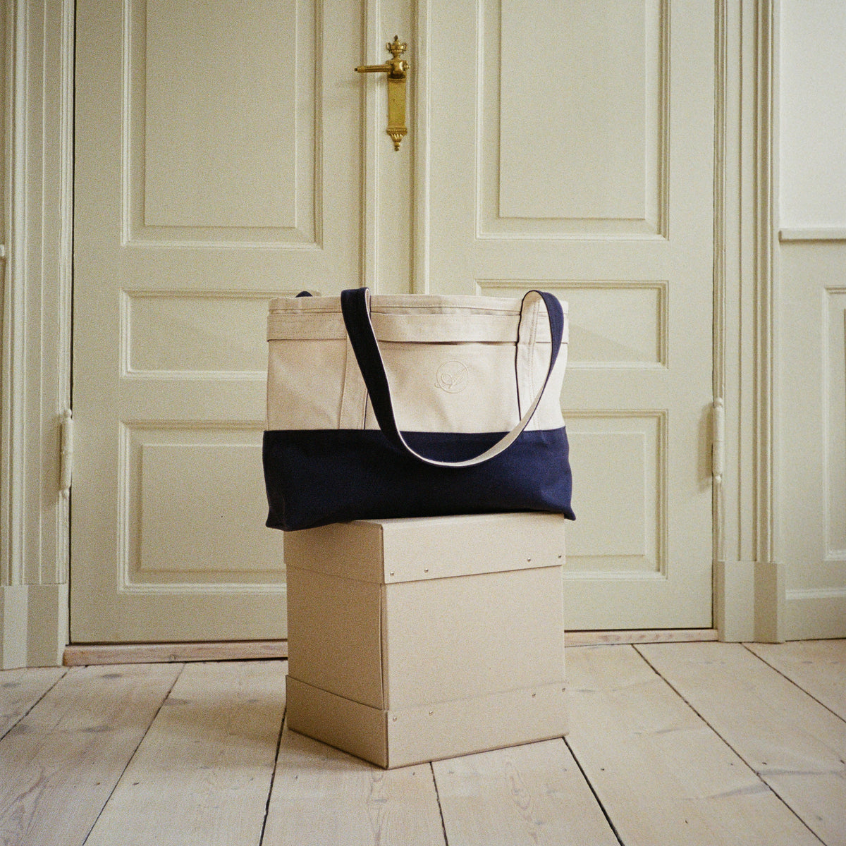Babette's Travel Bag in beige and navy on a pale box in front of white panelled doors, front view
