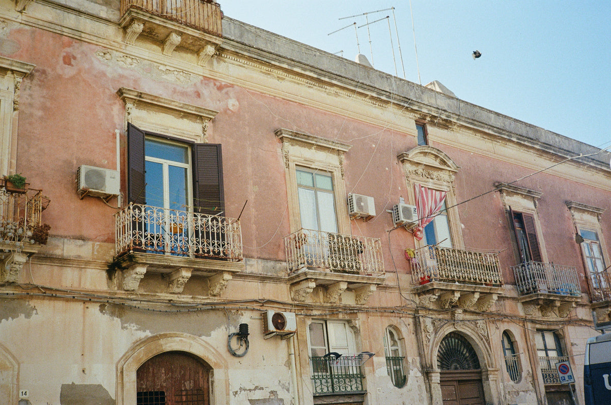 A weathered pink baroque facade with wrought-iron balconies in Siracusa