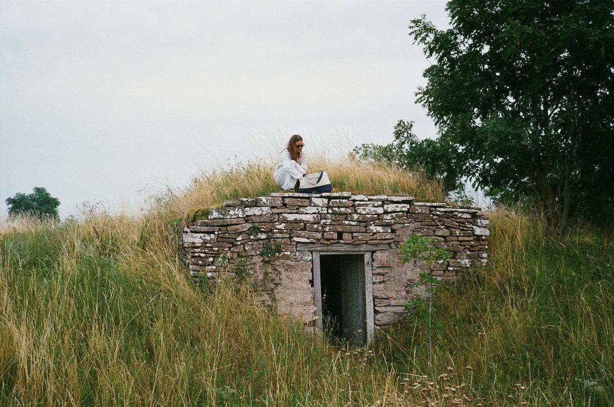 A woman with an Abelo tote sitting on an old stone root cellar in a summer meadow