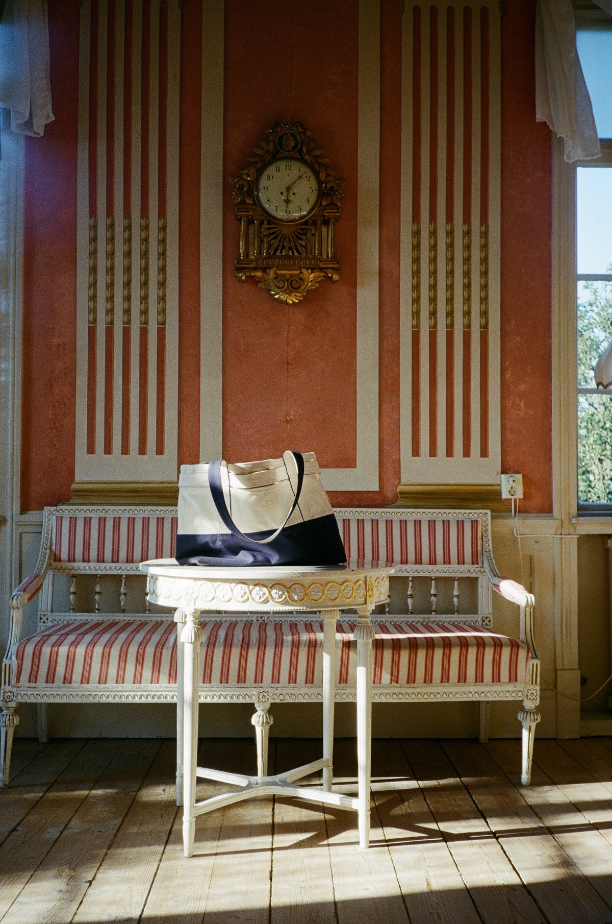 A cream and navy Abelo tote bag on an antique table in a terracotta room with a gilded wall clock