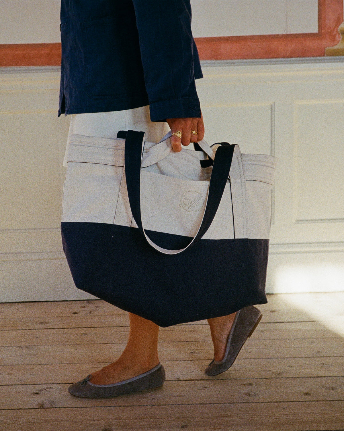 A woman carrying a cream and navy Abelo tote bag across a wooden floor